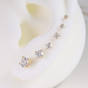 Peut inclure: Présentation de six boucles d'oreilles à clous graduées avec des pierres précieuses claires et étincelantes serties dans de l'or. Les boucles d'oreilles sont disposées en ligne, de taille décroissante, sur un modèle d'oreille blanc.