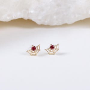 Może przedstawiać: Para złotych kolczyków w kształcie pajęczyny, każdy z małym, okrągłym, czerwonym kamieniem szlachetnym pośrodku. Kolczyki umieszczone są na białym tle z teksturowanym, kremowym kamieniem w górnej części obrazu.