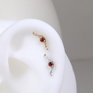 Pode incluir: Dois piercings de orelha ornamentados, um dourado e outro prateado, cada um com uma pequena pedra preciosa vermelha redonda. Os piercings têm um design delicado e ondulado. A joia é exibida em um modelo de orelha branca.