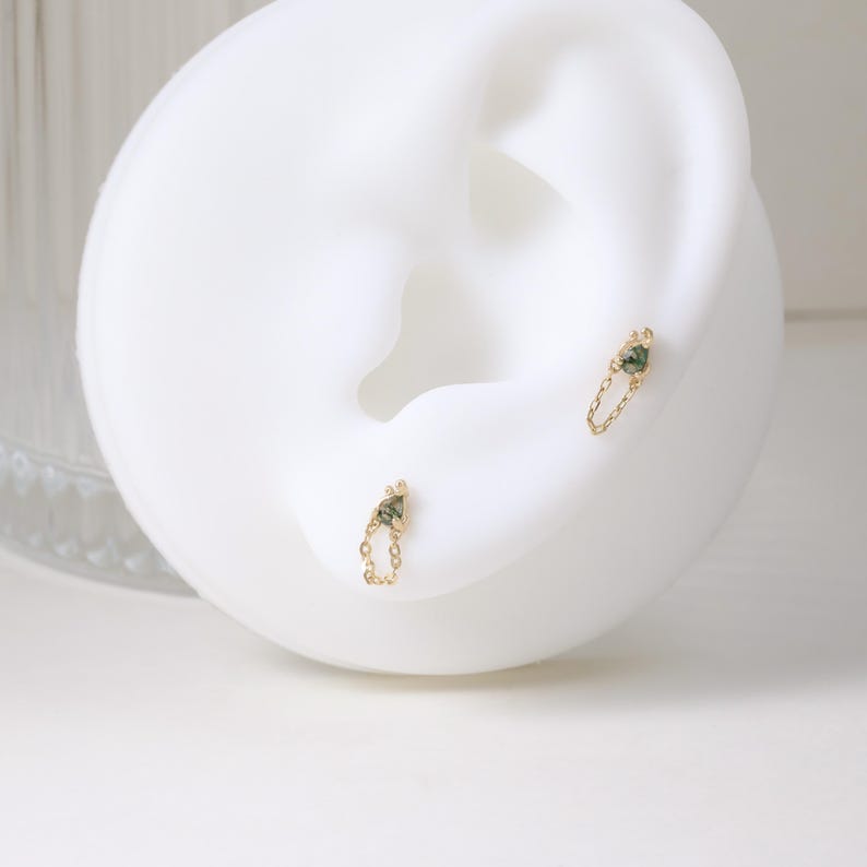 May include: A pair of gold earrings with a chain design. Each earring features a small, triangular green gemstone at the top and a delicate chain that dangles below. The earrings are displayed on a white ear-shaped form.