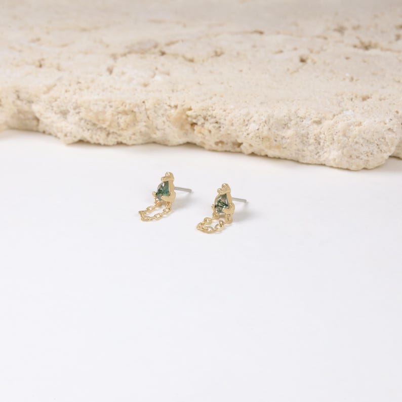 May include: A pair of gold-colored stud earrings with a unique design. Each earring features a small, irregularly shaped green stone and a delicate chain detail. The earrings are set against a white background with a textured, light-colored stone in the background.