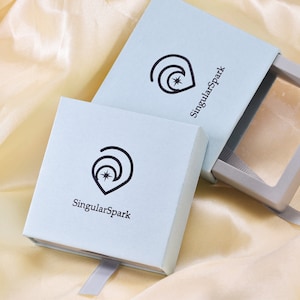 Puede incluir: Dos cajas de color azul claro con una cinta blanca. Las cajas tienen un logotipo con una luna creciente y una estrella, y el texto "SingularSpark".