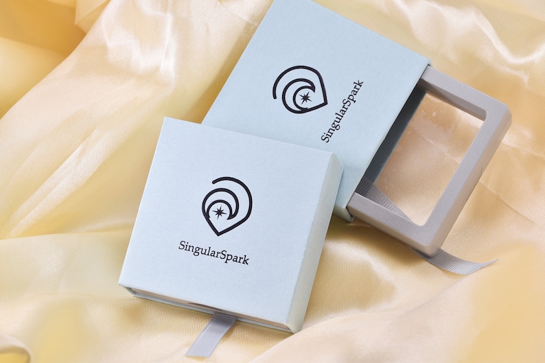 Puede incluir: Dos cajas de color azul claro con una cinta blanca. Las cajas tienen un logotipo con una luna creciente y una estrella, y el texto "SingularSpark".
