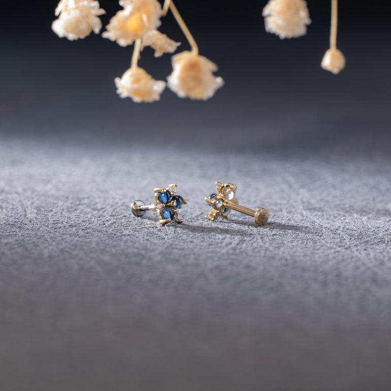 Puede incluir: Un par de piezas de joyer&iacute;a corporal de tono dorado con piedras preciosas azules. La joyer&iacute;a tiene forma de estrella con un peque&ntilde;o poste para perforar.