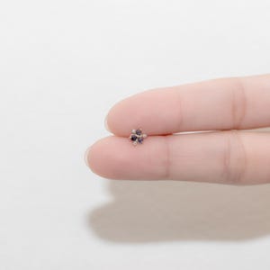Puede incluir: Un peque&ntilde;o pendiente de bot&oacute;n en forma de flor de color dorado con cinco piedras preciosas de color morado y blanco.