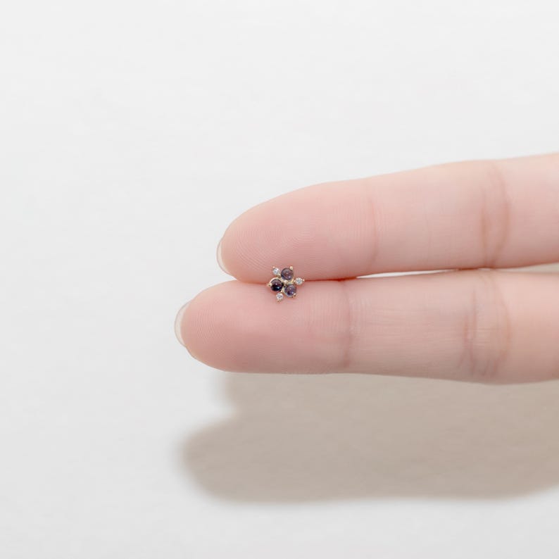 Puede incluir: Un peque&ntilde;o pendiente de bot&oacute;n en forma de flor de color dorado con cinco piedras preciosas de color morado y blanco.