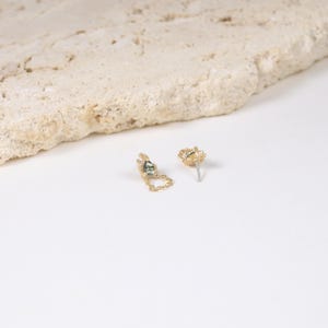 May include: A pair of gold-colored earrings. One earring features a small, square, green gemstone with a delicate chain. The other earring is a stud with a textured design. The earrings are displayed on a white surface.