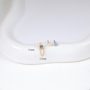 May include: A gold-colored earring with a chain and a small, green gemstone, measuring 11mm in length and 3.5mm in width. Two other small earrings are also present. The items are displayed on a white surface.