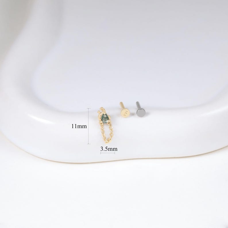 May include: A gold-colored earring with a chain and a small, green gemstone, measuring 11mm in length and 3.5mm in width. Two other small earrings are also present. The items are displayed on a white surface.