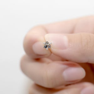 Puede incluir: Un pendiente de bot&oacute;n de oro en forma de flor con cinco piedras preciosas transparentes y negras.