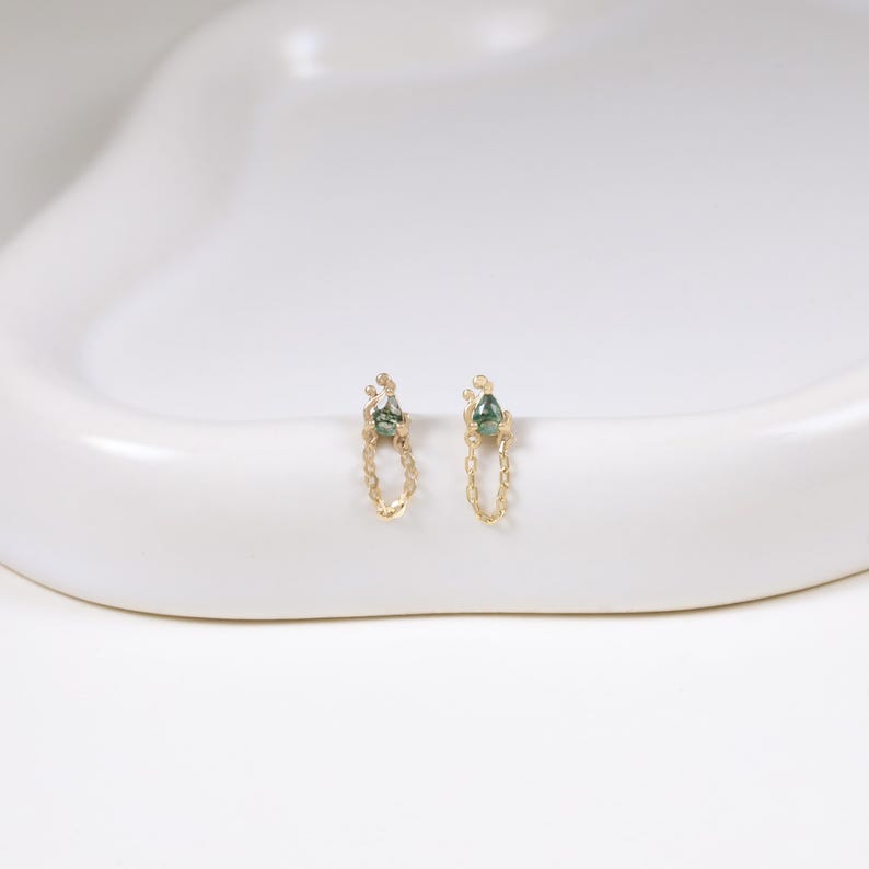 May include: A pair of gold chain earrings with a small green gemstone at the top and bottom. The earrings are displayed on a white, curved surface. The gemstones have a teardrop shape.