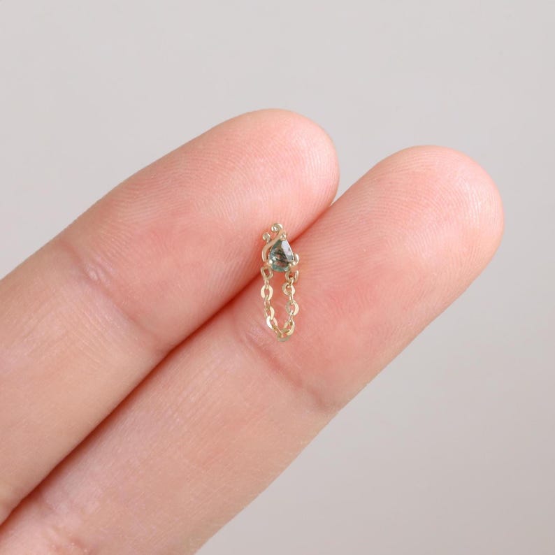 May include: A delicate gold-colored earring with a small, gray gemstone and a dangling chain. The earring is held between two fingers against a soft, neutral background. The jewelry piece is small and elegant.