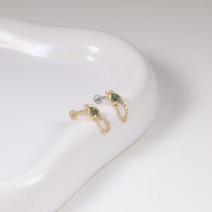 May include: Two gold-colored earrings with a small green gemstone and a delicate chain design. The earrings are displayed on a white surface, showcasing their intricate details and elegant style. The jewelry is designed for ear piercings.