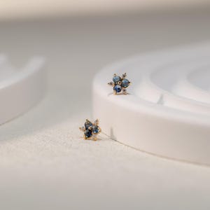 Puede incluir: Un par de pendientes de oro con un dise&ntilde;o de flor. Cada pendiente tiene cuatro piedras preciosas azules rodeadas de piedras preciosas transparentes m&aacute;s peque&ntilde;as.