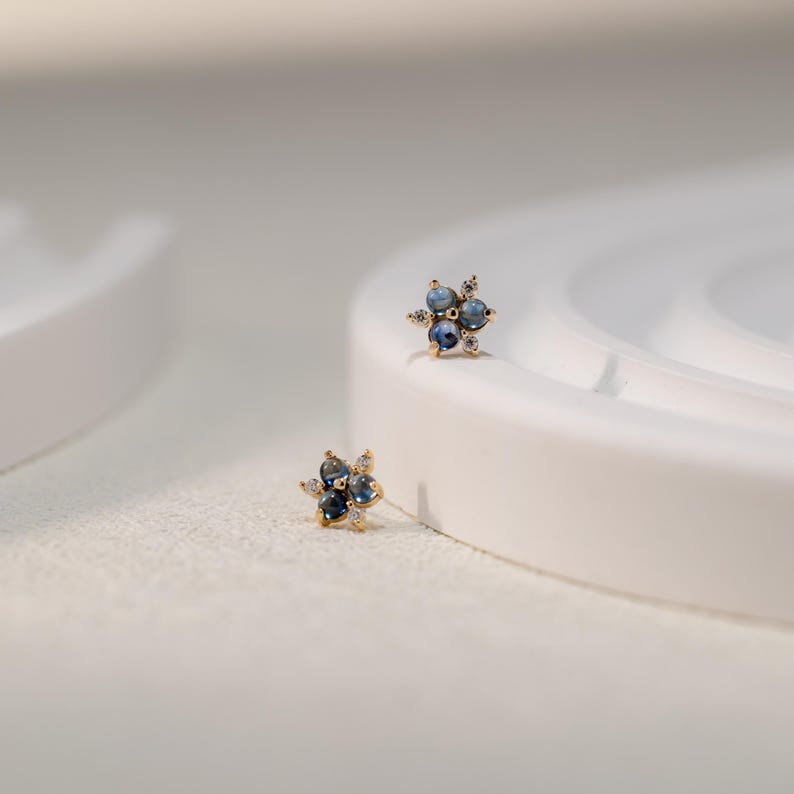 Puede incluir: Un par de pendientes de oro con un dise&ntilde;o de flor. Cada pendiente tiene cuatro piedras preciosas azules rodeadas de piedras preciosas transparentes m&aacute;s peque&ntilde;as.