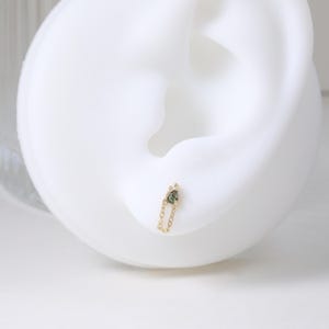 May include: A gold chain earring with a small, dark green gemstone. The earring is attached to a white ear model. The chain is delicate and the gemstone is set in a simple, elegant design. The earring is a piece of jewelry.