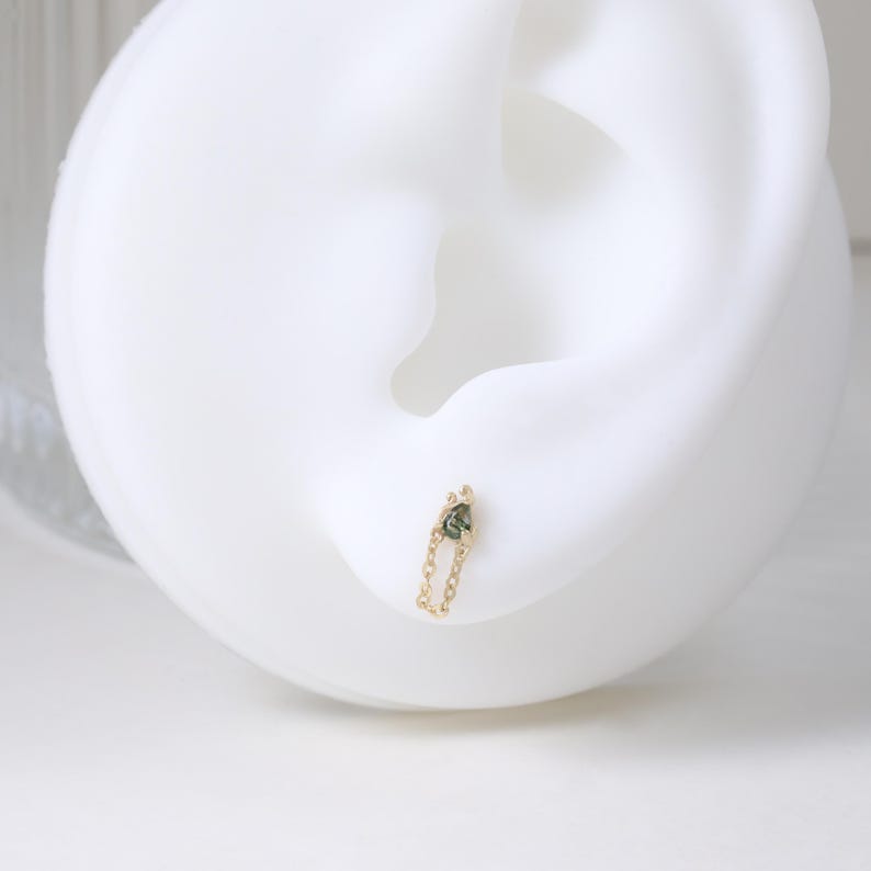 May include: A gold chain earring with a small, dark green gemstone. The earring is attached to a white ear model. The chain is delicate and the gemstone is set in a simple, elegant design. The earring is a piece of jewelry.
