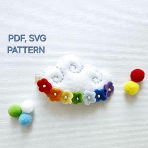 Op de afbeelding: Een witte wolkenvorm van vilt met regenboogkleurige viltbloemen en witte parelcentra. De wolk heeft een witte vilt achterkant en een wit lint. De tekst "PDF, SVG PATTERN" staat boven de wolk.