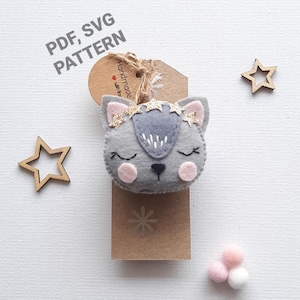 Op de afbeelding: Een grijze viltkat ornament met roze wangen en gouden glittersterren. De ornament is bevestigd aan een bruine kaart met de tekst "Handmade with love" en "PDF, SVG PATTERN".