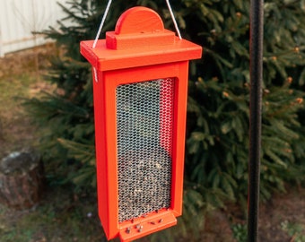 Op de afbeelding: Een rode vogelvoederhuisje met een metalen gaas aan de voorkant hangt aan een zwarte metalen haak. Het voederhuisje is gevuld met vogelzaad en is omgeven door groen gebladerte.
