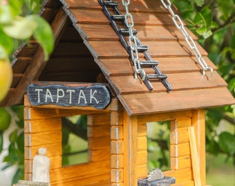 Op de afbeelding: Een houten vogelvoederhuisje in de vorm van een klein huis met een bruin dak, een kleine ladder en een bord met de tekst "TAPTAK". De voederbak hangt aan een ketting en is omgeven door groene bladeren.