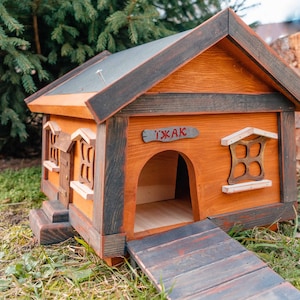 Puede incluir: Una caseta para perros de madera con una rampa, pintada en tonos marrones y negros. La casa tiene una pequeña ventana y un letrero que dice "T'AK".