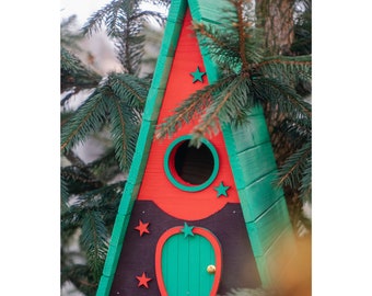 May include: A triangular birdhouse painted in red, green, and black with a small green door and a round opening. The birdhouse has a brown base and is decorated with stars.