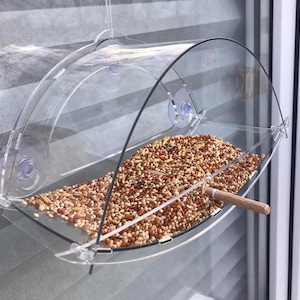 Puede incluir: Un comedero para pájaros de plástico transparente con un techo curvo y ventosas. El comedero está lleno de una mezcla de semillas para pájaros.
