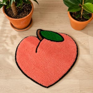 May include: A peach-shaped rug with a black outline, a green leaf, and a brown stem. The rug is a soft, textured material in a peachy-pink color. Two potted plants are in the background.