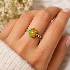 Könnte beinhalten: Ein Silberring mit einem ovalen Opal-Edelstein, der lebhafte orange, gelbe und grüne Farbreflexe aufweist. Der Stein ist in einer Vier-Krappen-Fassung auf einer glatten, polierten Silberschiene gefasst, die den Finger mit einem schlichten Design umschließt.