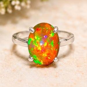 Könnte beinhalten: Ein Silberring mit einem großen, ovalen Cabochon-Opal. Der Opal zeigt ein lebhaftes Farbspiel mit intensiven orangefarbenen, roten und hellgrünen Blitzen im gesamten Stein. Der Edelstein wird von vier silbernen Krappen sicher gehalten.