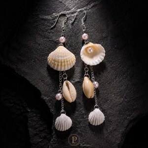 Könnte beinhalten: Hängeohrringe mit silberfarbenen Haken, feinen Ketten, kleinen rosa Perlen und verschiedenen natürlichen Muscheln. Das Design umfasst jeweils eine größere gerippte Muschel, eine glatte Kaurimuschel und eine kleine weiße fächerförmige Muschel.