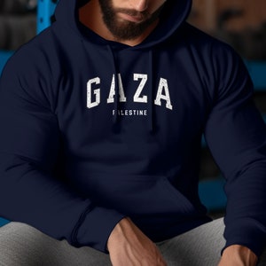 Puede incluir: Una sudadera con capucha azul marino con un estampado gráfico blanco que dice "GAZA PALESTINE" en una fuente de estilo vintage.
