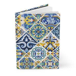 Può includere: Un diario con copertina rigida con un colorato motivo a piastrelle. Il design presenta un motivo ripetuto di forme geometriche blu, gialle e bianche. Il diario è aperto, rivelando pagine bianche.