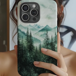 Può includere: Custodia per smartphone con un dipinto ad acquerello di un paesaggio di foresta e montagna. Il design presenta alberi sempreverdi in primo piano e montagne nebbiose sullo sfondo, con sfumature di verde e blu. La custodia ha un ritaglio per la fotocamera.
