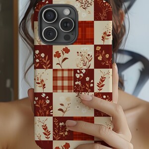 May include: A phone case with a patchwork design featuring floral and plaid patterns in shades of red, cream, and brown. The case has a camera module with three lenses and a flash.