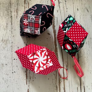Peut inclure: Trois décorations de Noël faites à la main. L'une est noire avec des motifs de cannes en sucre et de tartan. Une autre est rouge avec des pois blancs et un motif de menthe poivrée. La troisième est rouge avec des pois blancs et le texte "Peace" et "Merry". Chacune a une boucle de ruban rouge.