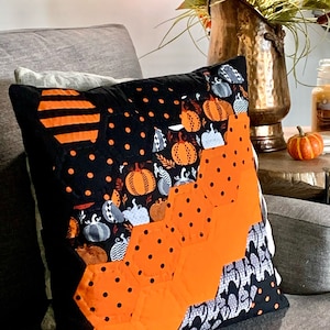 Könnte beinhalten: Ein orange-schwarzes Halloween-Kissen mit einem geometrischen Muster aus Hexagonen und Kürbissen. Das Kissen liegt auf einem grauen Sofa.