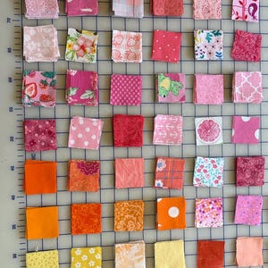 May include: A grid of fabric squares in various shades of pink, orange, and yellow. Each square features a unique pattern, including florals, polka dots, and geometric designs. The squares are arranged on a gray surface with a grid pattern.