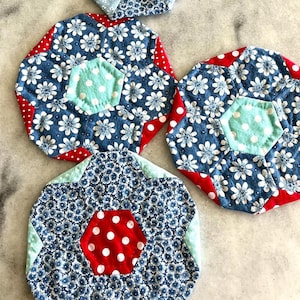 Może przedstawiać: Kolekcja ręcznie robionych podkładek z tkaniny. Każda podkładka ma niebieski kwiatowy wzór z białymi kwiatami, czerwoną obwódkę w kropki i centralny sześciokąt w kolorze czerwonym z białymi kropkami lub miętowym z białymi kropkami.