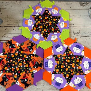 Pode incluir: Três decorações de tecido com tema de Halloween. Cada uma tem um centro em forma de estrela com fundo preto e doces de milho. As bordas externas são roxas, laranja e verdes. Uma apresenta gatos pretos, outra cupcakes e a terceira, cupcakes.