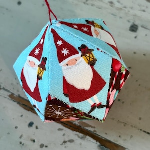 May include: A fabric Christmas ornament with a geometric shape. The ornament is covered in light blue fabric printed with Santa Claus figures, snowflakes, and other holiday designs. A red string is attached for hanging.