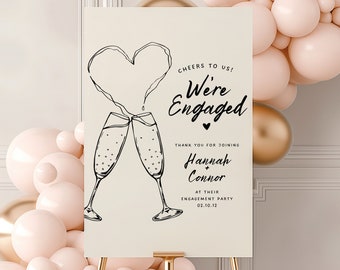May include: A white sign with black text and a hand-drawn illustration of two champagne glasses and a heart. The text reads "Cheers to us! We're Engaged" and "Thank you for joining Hannah + Connor at their engagement party 02.10.12".