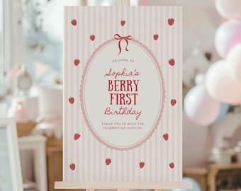 May include: A welcome sign on a wooden easel with a pink and white striped background. The sign reads "Welcome to Sophia's BERRY FIRST Birthday" with strawberry illustrations. A red bow and strawberries decorate the sign.