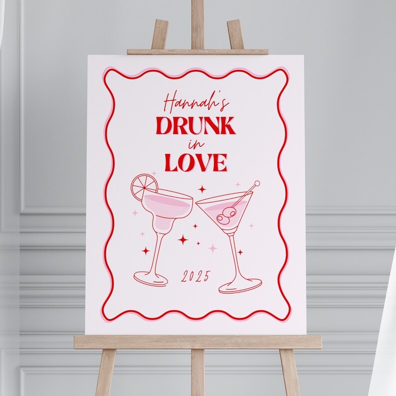 May include: A white sign with pink and red borders and text that says "Hannah's Drunk in Love 2025". Two pink cocktail glasses with a lime wedge and olives are illustrated on the sign.