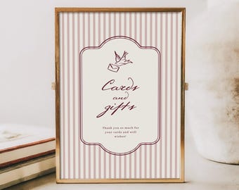 Pode incluir: Uma placa emoldurada com uma moldura de metal dourado. A placa tem um fundo listrado vertical em rosa claro e branco. O centro apresenta um rótulo decorativo com as palavras "Cards and gifts" em uma fonte de script e uma pequena ilustração de um pássaro.
