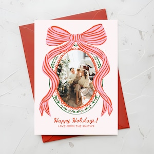 May include: A holiday greeting card with a family photo inside an oval frame, surrounded by a green wreath and a pink bow with red stripes. The card reads "Happy Holidays! LOVE FROM THE SMITH'S" and is set against a red envelope.