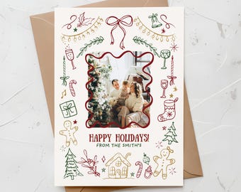 Könnte beinhalten: Eine Weihnachtsgrußkarte mit einem Familienfoto, das von einem roten, wellenförmigen Rand umrahmt ist. Die Karte zeigt festliche Kritzeleien in Grün und Gold, darunter eine Schleife, Lichter und einen Lebkuchenmann. Der Text lautet "HAPPY HOLIDAYS! FROM THE SMITH'S". Die Karte befindet sich in einem braunen Umschlag.