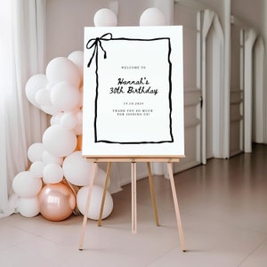 K&ouml;nnte beinhalten: Ein wei&szlig;es Willkommens-Schild mit schwarzem Rand und einer schwarzen Schleife oben. Das Schild lautet "Willkommen zu Hannah's 30th Birthday 19.10.2024 Vielen Dank, dass Sie mit uns feiern!"
