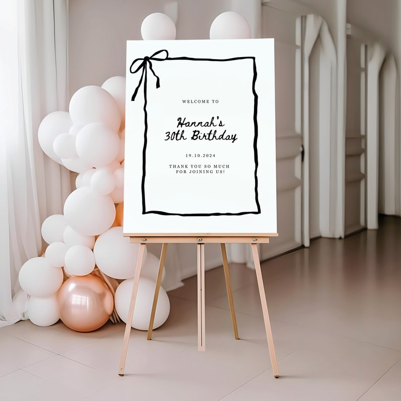 K&ouml;nnte beinhalten: Ein wei&szlig;es Willkommens-Schild mit schwarzem Rand und einer schwarzen Schleife oben. Das Schild lautet "Willkommen zu Hannah's 30th Birthday 19.10.2024 Vielen Dank, dass Sie mit uns feiern!"